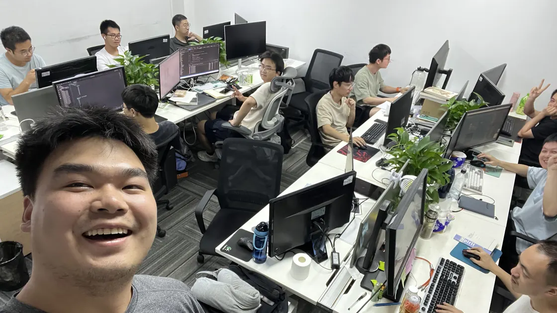 Selfie of Junhua in the Farsee2 office, smiling in the foreground, with colleagues at their desks and monitors behind him.